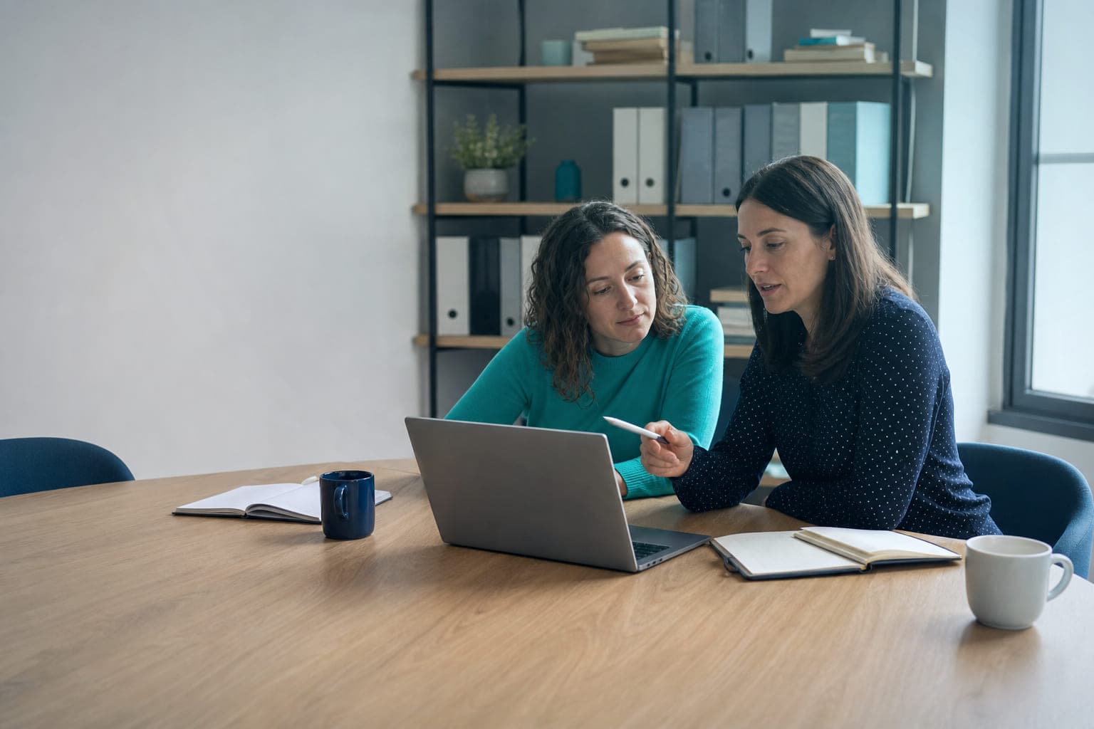 Dos compañeras de una asesoría revisan juntas la pantalla de un portátil en la mesa de reuniones, con cuadernos y café al lado.