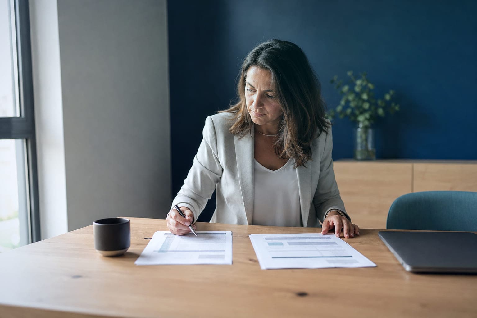 Una mujer compara dos propuestas impresas, una a cada lado, sobre la mesa de madera de una oficina pequeña, con el bolígrafo en la mano y el portátil cerrado al lado.