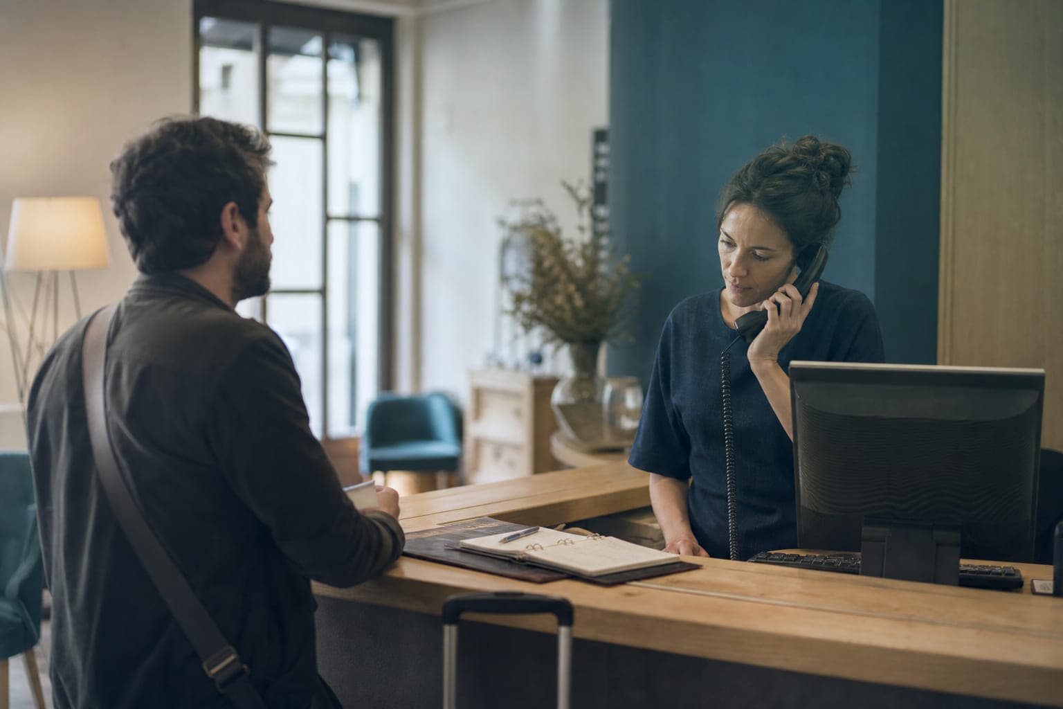 Una recepcionista de hotel sostiene el teléfono con gesto de paciencia mientras atiende a un huésped que espera al otro lado del mostrador.