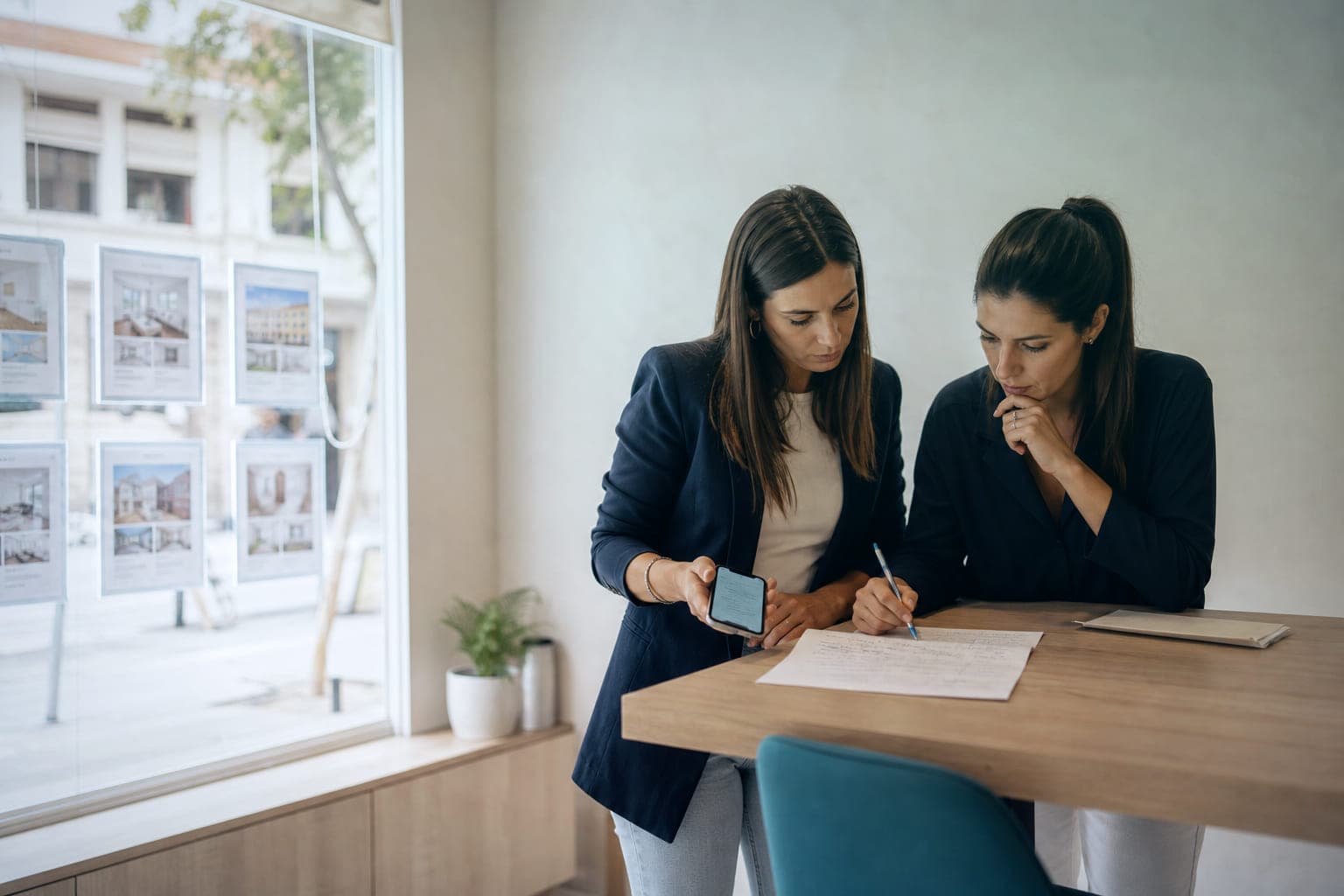 Dos comerciales de una agencia inmobiliaria, de pie junto a una mesa alta, repasan una hoja con anotaciones: una sostiene el móvil con una conversación abierta y la otra escribe con un bolígrafo. En el escaparate, fichas de pisos.