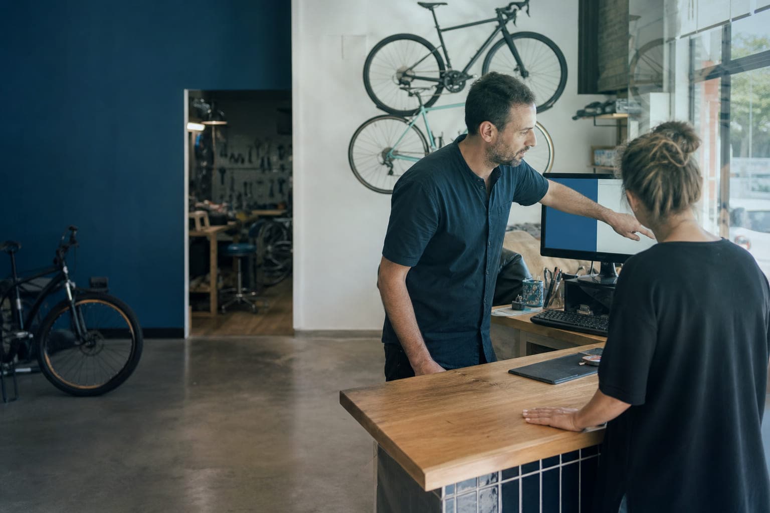 El dueño de una tienda de bicicletas atiende a una clienta en el mostrador mientras consulta la ficha de un pedido en el ordenador.